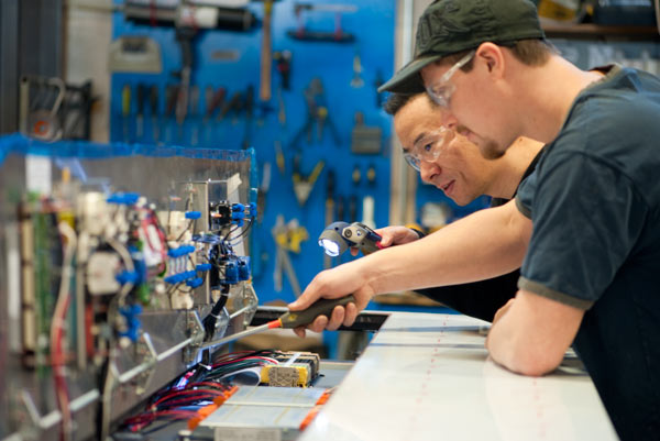 Two technicians working on a machine, one holding a screwdriver the other a flashlight